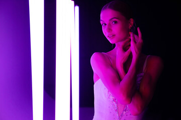 Portrait of a ballerina in the role of a white swan against a background of bright neon lights in a dark studio. Beautiful young model gracefully posing with her hands.