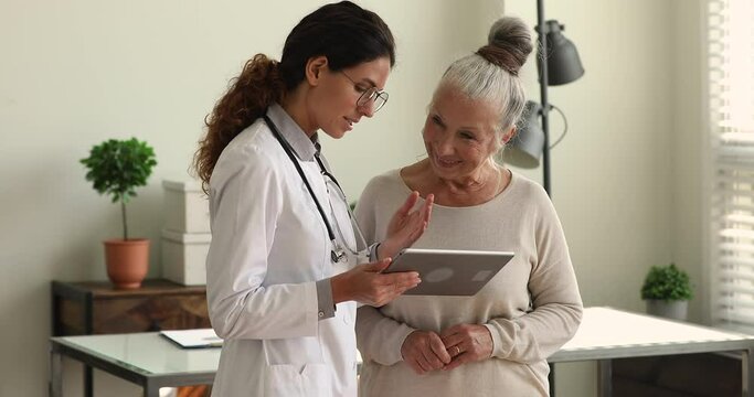 During visit young female doctor in white coat showing to older female patient health test result, explain prescription using medical app on digital wireless tablet. Elder care, modern tech concept