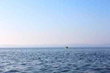 Fototapeta premium Man in a green kayak paddle at the horizon of blue lake in autumn season