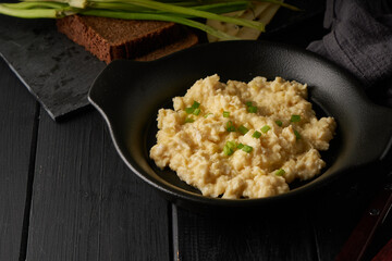 Scrambled eggs with green onion served in dark plate with bread on table