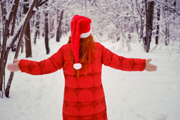 Woman stands with her back to the ground her arms outstretched among the winter trees in the snow. Red-haired woman on Christmas and New Year Eve walks in the winter in red clothes