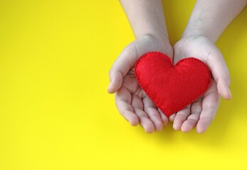 Red heart in the hands of a child on a yellow background with copy space. The concept of helping children, hope and love.