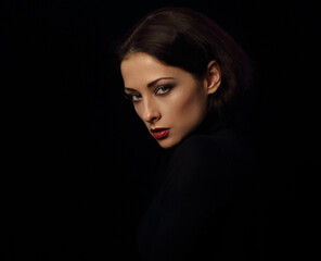 Sexy vamp woman with bright evening makeup, red lipstick brown brow posing isolated on dark black background. Closeup portrait in deep low key light shadows.