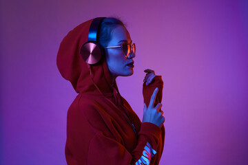 Fashion hipster woman wear stylish glasses and headphones listening to music over color neon background at studio.
