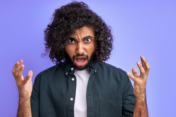 disgruntled curly male shout, spread arms, he is irritated by news or someone's behavior, isolated on purple background