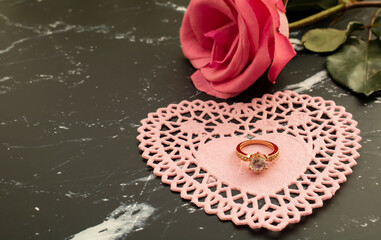 Single pink rose with a diamond ring on a pink heart doily. Black marble background. 