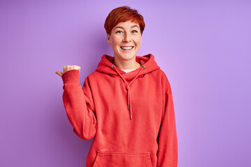 redhead woman showing at side, pointing finger and smiling, isolated on purple background