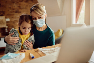 Mother with face mask using laptop while taking care of her ill daughter at home.