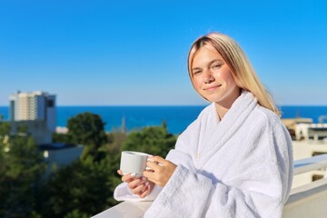 Happy young woman in bathrobe enjoying cup of coffee and sunny landscape of sea resort