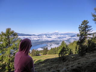 A woman in an orange hoodie enjoying the idyllic view on Alpine Millstaettersee lake in Austria. She is hiking in high Alps. The valley is shrouded with some clouds, sunny day. The girl is calm, happy