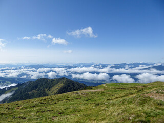 A panoramic view on the Alpine valley from the top of Granattor in Austrian Alps. The valley is shrouded in fog. The slopes are lush green. Many mountain poking above the clouds. Mystery mood