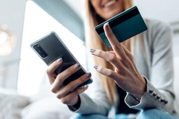 Smiling young woman paying something online with her credit card and the smartphone while sitting on sofa at home.