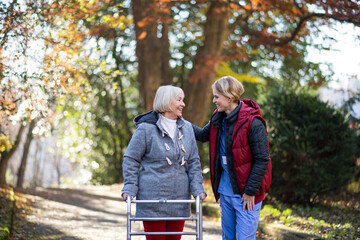 Senior woman with walking frame and caregiver outdoors on a walk in park, coronavirus concept.