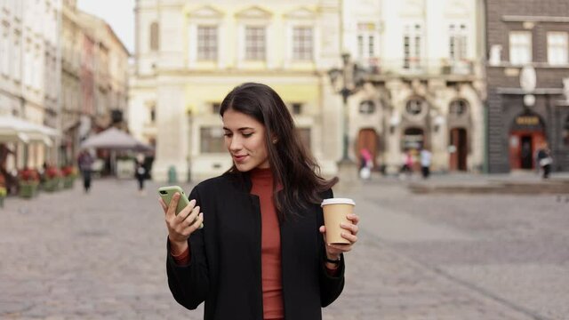 Beautiful young woman on vacation walking around the city, enjoying the sights, checks online maps on smartphone, drinking coffee. Tourism, travelling, rest, relaxation