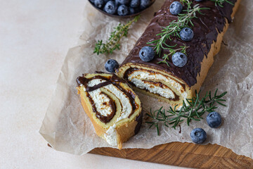 Sponge cake roll with chocolate and cream cheese decorated with chocolate glaze, blueberry and rosemary on parchment, light concrete background. Biscuit swiss roll.