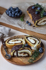 Sponge cake roll with chocolate and cream cheese decorated with chocolate glaze, blueberry and rosemary on parchment, light concrete background. Biscuit swiss roll.