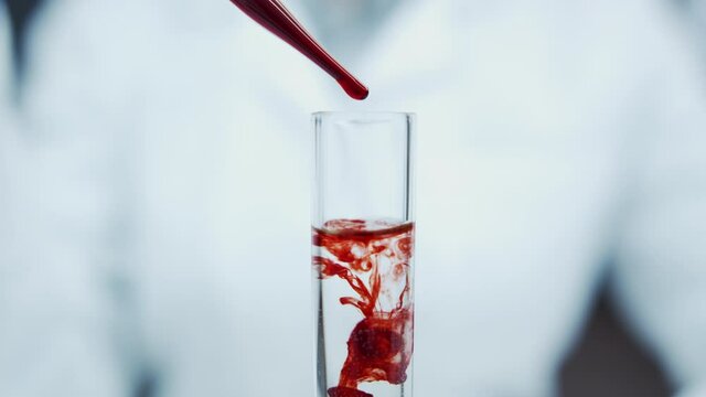 Close-up of pipette tube in the hands of doctor. Blood drips from the pipette. Blood sampling for tests. Creation of vaccine against infections. Blood drops dissolve in clear liquid in test tube.
