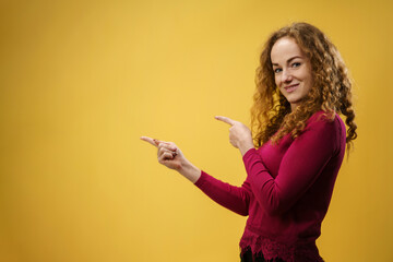 Portrait of young woman in a studio on yellow background, pointing fingers to copy space.