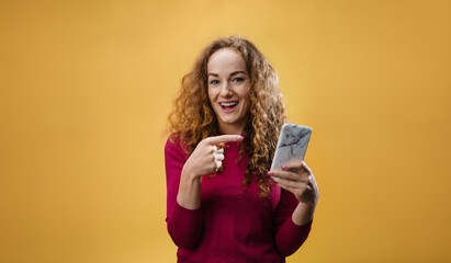 Portrait of young woman in a studio on yellow background, pointing fingers to smartphone.