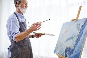 artist man is drawing on easel canvas, senior gray haired man in apron enjoy the process of painting, in light room. side view