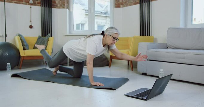 Beautiful active 65-aged grey-haired woman in white t-shirt and leggins doingstretching exercises on mat at home while watching video online on computer