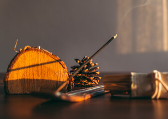 Fototapeta na wymiar Burning incense stick in a scene natural and wooden decoration objects during sunset. Selective focus on stick tip ash. Cozy home environment. Warm toned photo.