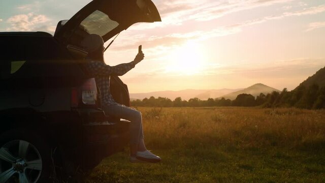 woman traveler holding smartphone taking selfie photo sitting car trunk looking on beautiful mountain landscape summer sky at sunset