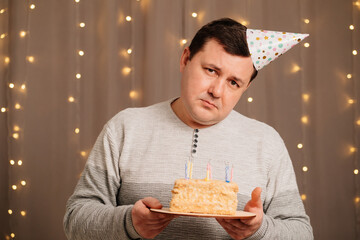 sad man in a festive cap with a birthday cake blows out candles. sadness due to aging and birthday....