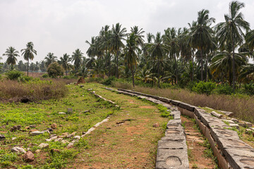 Fototapeta premium Hampi, Karnataka, India - November 5, 2013: Ancient Aqueduct. flows through green landscape with palm trees under silver sky. Grass and weeds on surface. Gray stone borders.