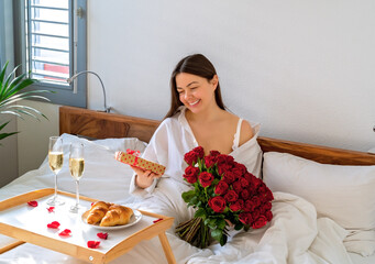 Happy woman having surprise gift and romantic breakfast in bed on Valentine Day holding present and...
