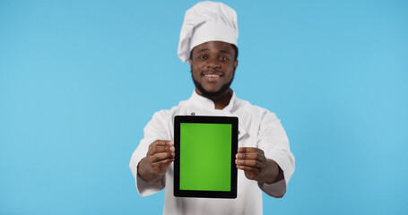 Portrait of happy African american chef cook in uniform showing digital tablet with blank screen standing isolated over blue background.