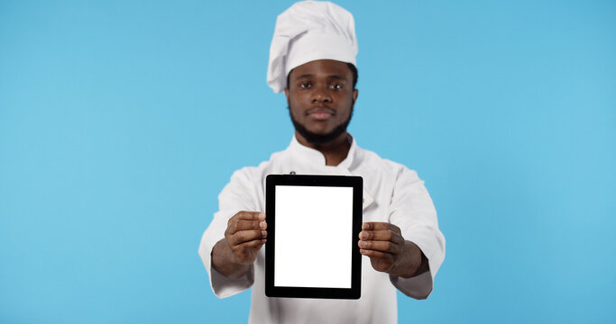 Portrait of African american chef cook in uniform showing digital tablet with blank screen standing isolated over blue background.
