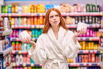 thoughtful female is choosing the best toilet paper in store, wearing white bathrobe, stand having fun in the household goods department in supermarket