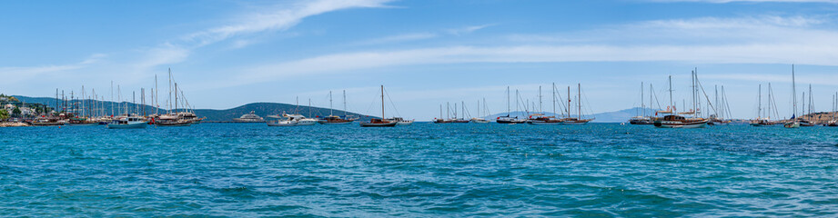 Fototapeta premium Panoramic distant view at yachts and sail boats anchored in calm waters of Bodrum shallow moon-shaped bay in Turkey.