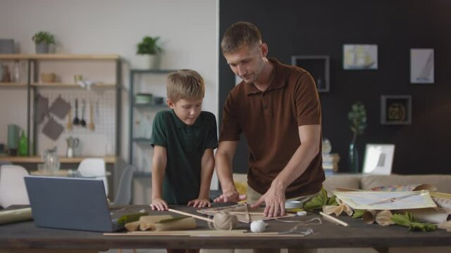 Medium shot of father and son spending time together crafting bright flying paper kite standing at table in living room