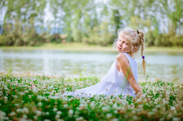 Fototapeta na wymiar fashionable child in white dress sitting in grass by the river and posing in summer time