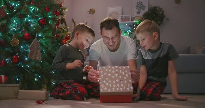 father with two children opens a Christmas present under the tree and rejoices. A gift for my father. A ray of light from the gift shines on the face. Rejoice with the children New Year's gift.