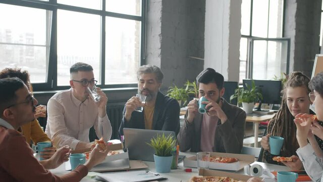 Happy office workers men and women are eating pizza during work break talking enjoying food sitting at table together. Business lunch and employees concept.