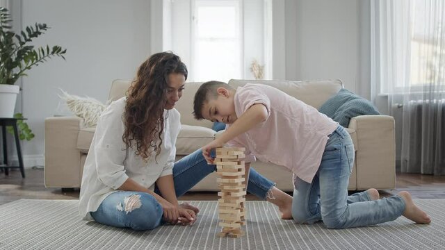Mother And Son Sitting On The Floor In The Apartment. They Play Game Of Assembling Pyramid Of Wooden Bars.