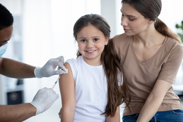 Cute happy girl visiting doctor with her mom, getting injection of covid-19 vaccine at hospital