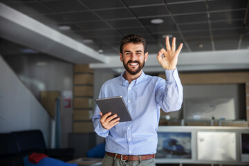 Smiling bearded CEO standing in lobby of export firm, showing okay gesture and holding tablet....