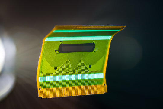 Green flexible circuit board floating on black background with white light rays. Closeup of miniature bent plastic flex PCB for data signals parallel transmission in LCD screens of electronic devices.