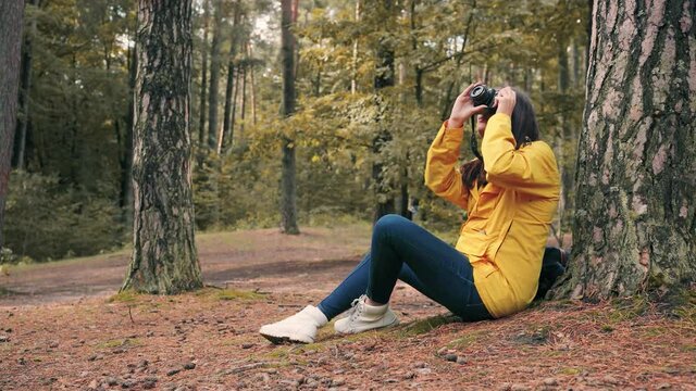 Young beautiful woman tourist in yellow jacket sits with backpack in woods under tree and takes photos of forest on camera.