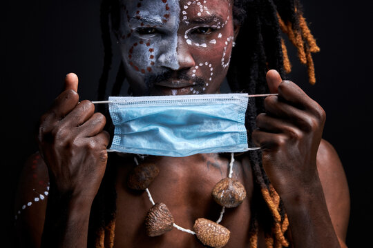 Aborigen Male Wearing Medical Mask On Face During Coronavirus, Covid-19 All Over The World. Man With Shamanic Paintings On Face Looks At Camera Isolated On Black Background