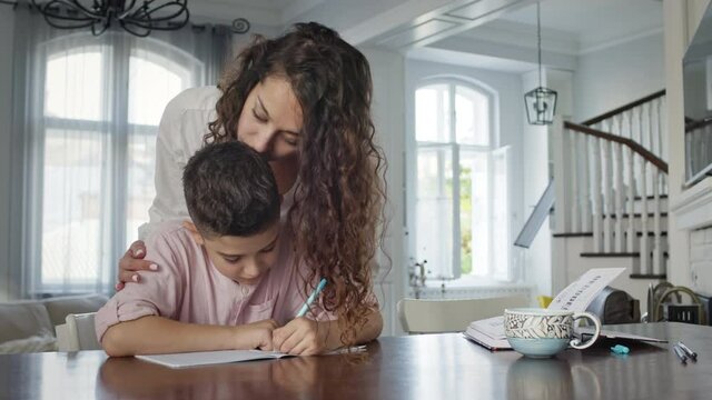Young Mother And Son At The Table. Boy Writes In Notebook. He Does His Homework.