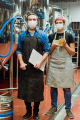 Two young brewers in workwear and protective masks standing in front of camera