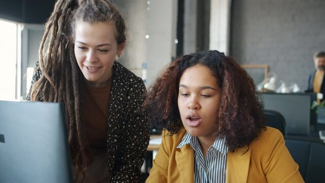 Cheerful girls Caucasian and Afro-American are talking using computer smiling laughing in workplace. Businesswomen and communication concept.