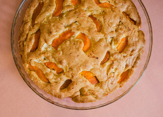 A large, round baked apple and apricot pie stands in a glass form on a table with a tablecloth.