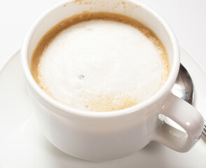 a white cup of coffee with creamy froth on the top on a saucer stands on a white table.