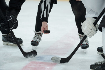 Hand of referee holding puck over ice rink and two hockey players with sticks